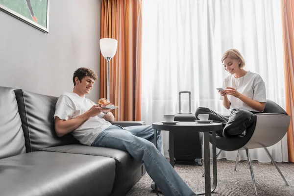Smiling woman using smartphone in armchair near boyfriend with croissant on couch in hotel room — Stockfoto