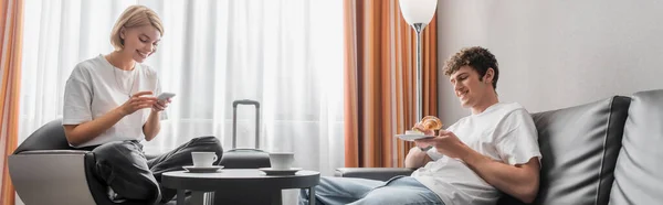Посмішка в кріслі за допомогою смартфона біля хлопця сидить з круасантом в номері готелю, банер — стокове фото