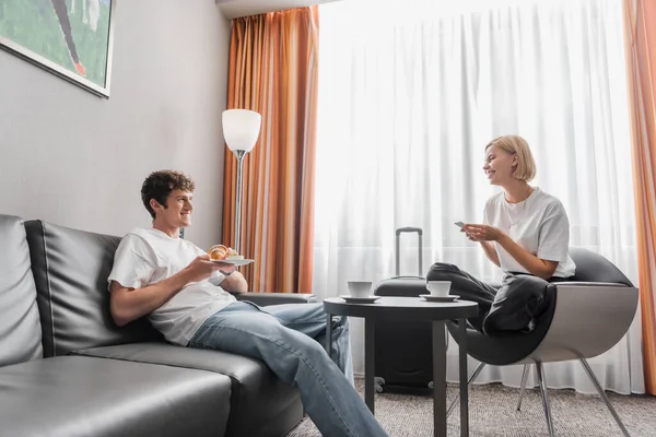 Young woman with smartphone sitting in armchair near boyfriend with croissant in modern hotel suite — Stockfoto