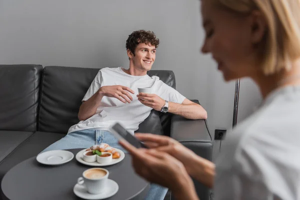 Усміхнений чоловік з чашкою кави сидить на дивані біля сніданку і розмита дівчина зі смартфоном у готельному номері — стокове фото