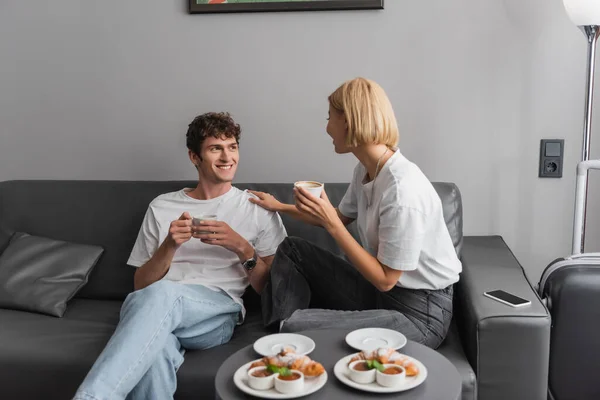 Blonde woman with cup talking to smiling boyfriend near croissants and jam with chocolate paste on coffee table - foto de stock