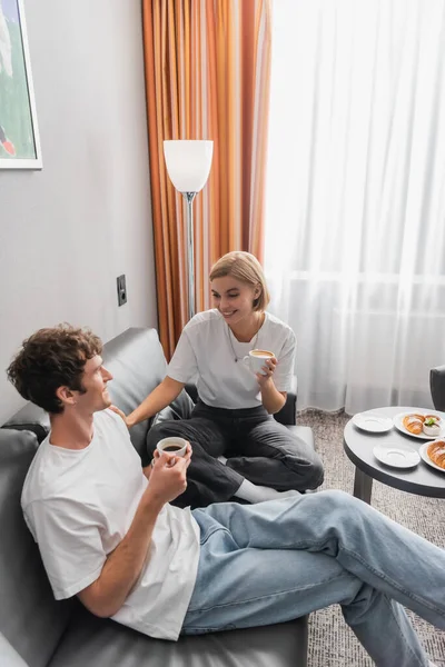 Веселая женщина с чашкой кофе разговаривает с парнем на диване возле завтрака в номере отеля — стоковое фото