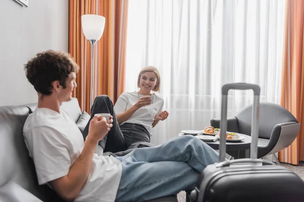 Happy couple of travelers drinking morning coffee near blurred suitcase in modern hotel suite — Stockfoto