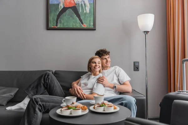 Веселая пара, обнимающаяся на диване возле вкусных круассанов и чашек кофе в отеле — стоковое фото