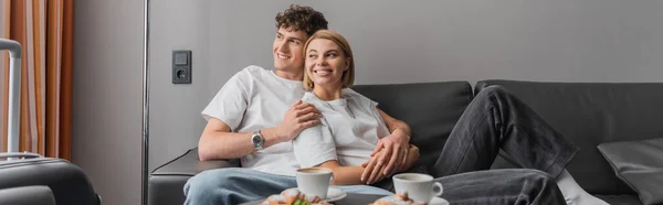 Радостная пара, обнимающая на диване возле кофейных чашек и смотрящая в отель, баннер — стоковое фото