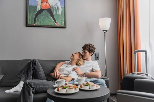 Happy young couple hugging on couch near cups and delicious croissants in hotel room — Stockfoto