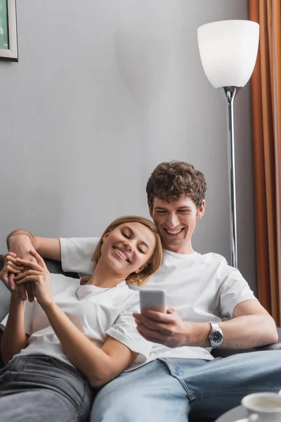 Pleased man using mobile phone and holding hands with happy girlfriend while resting in hotel apartments — Photo de stock