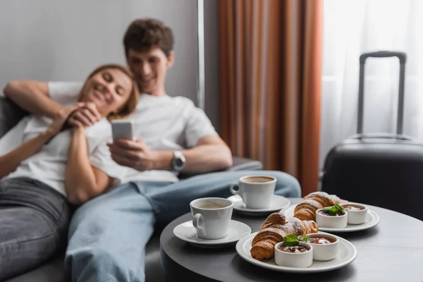 Coffee cups and croissants with jam and chocolate paste near blurred couple with smartphone in hotel room — Stockfoto