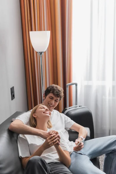 Счастливый мужчина со смартфоном, держащийся за руки с улыбчивой девушкой на диване в номере отеля — стоковое фото