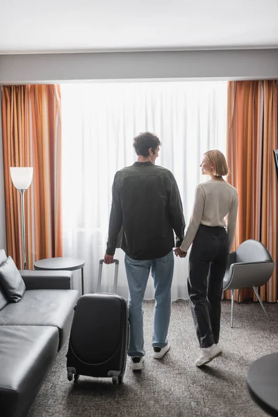 Back view of young tourists with suitcase holding hands and looking at each other in hotel room — Photo de stock