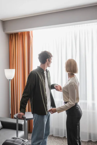 Young man with suitcase holding hands with girlfriend near window with curtains in hotel — Foto stock