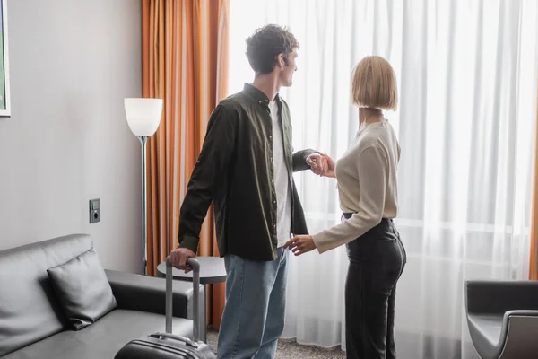 Couple of young tourists with travel bag holding hands near window with curtains in hotel suite — Foto stock