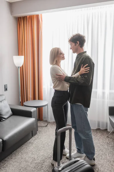 Full length of young couple hugging and looking at each other near travel bag in hotel room — Photo de stock