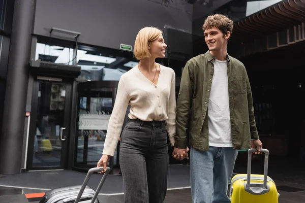 Couple of happy tourists with suitcases holding hands and looking at each other in hall of modern hotel — Stock Photo