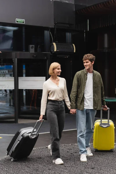 Щаслива молода пара тримає руки під час прогулянки з валізами в готельному лобі — стокове фото