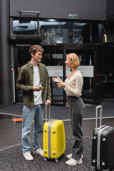 Young man with mobile phone smiling at blonde girlfriend talking near travel bags in hotel lobby - foto de stock