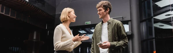 Cheerful blonde woman talking to smiling boyfriend holding mobile phone in hotel hall, banner — Stock Photo