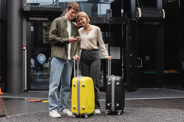 Full length of young man showing smartphone to happy blonde girlfriend near suitcases in lobby of hotel — Stockfoto