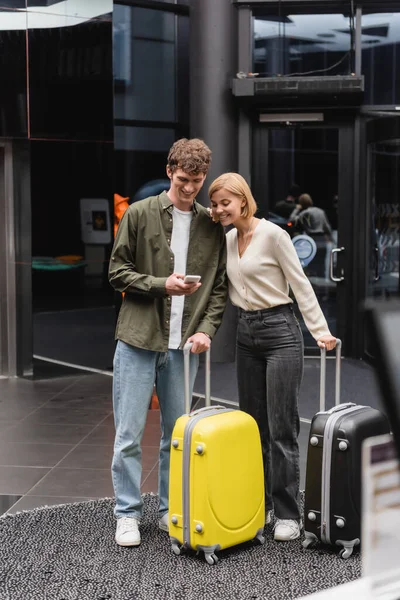 Happy young couple with travel bags looking at mobile phone in hall of modern hotel — Fotografia de Stock