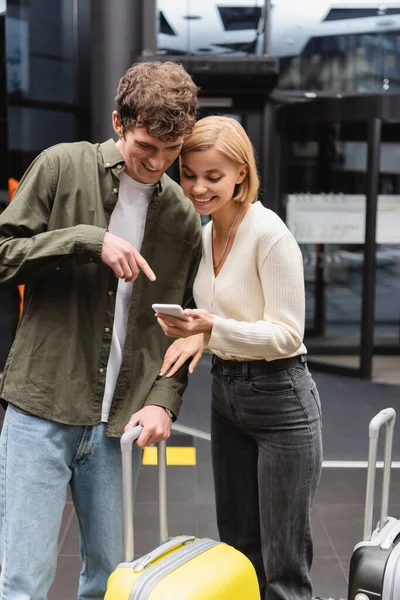 Улыбающийся мужчина указывает на мобильный телефон в руке веселой подруги возле чемоданов в отеле — стоковое фото