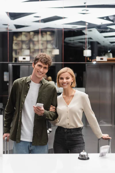 Blonde woman and brunette man with smartphone smiling at camera near reception in modern hotel — Stock Photo