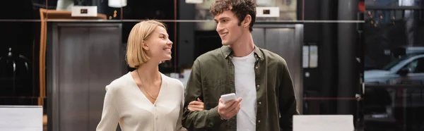 Cheerful blonde woman and man with mobile phone looking at each other in hotel lobby, banner — Stock Photo