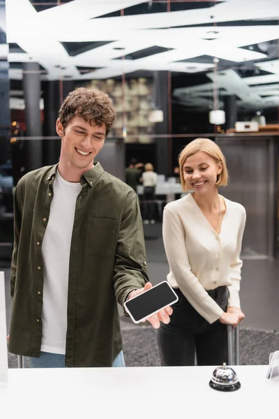 Happy man holding smartphone with blank screen near reception and girlfriend in hotel — Photo de stock