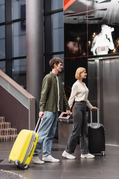 Side view of young couple with travel bags holding hands and walking in modern hotel - foto de stock