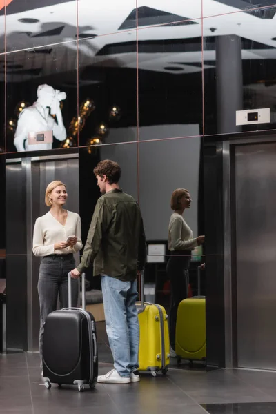 Young blonde woman with smartphone smiling near boyfriend with suitcases in lobby of modern hotel — Fotografia de Stock