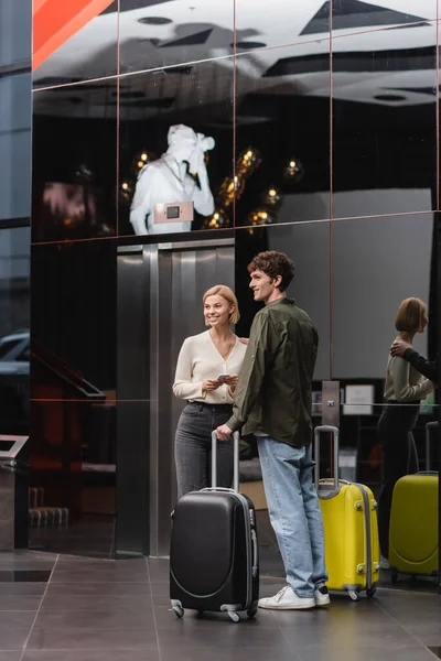 Smiling couple with suitcases looking away while standing with travel bags in modern hotel — Photo de stock