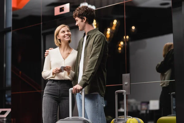 Blonde woman with cellphone smiling at young boyfriend near luggage in modern hotel — Stock Photo