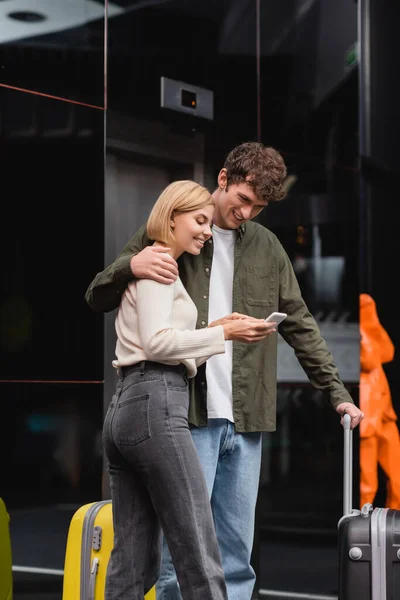 Улыбающаяся женщина показывает мобильный телефон своему парню, обнимающему ее возле багажа в вестибюле отеля — стоковое фото