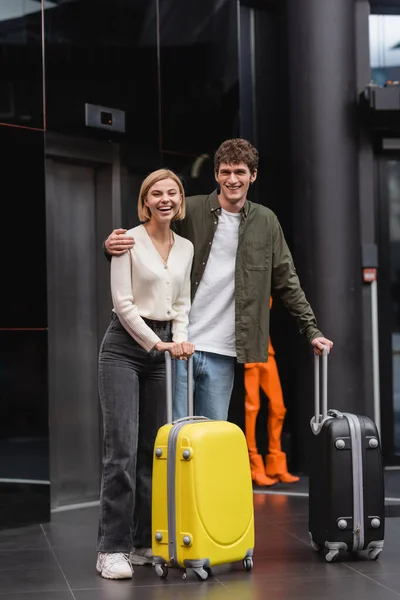 Молодой человек с блондинкой, стоящий с чемоданами и смеющийся в холле отеля — стоковое фото
