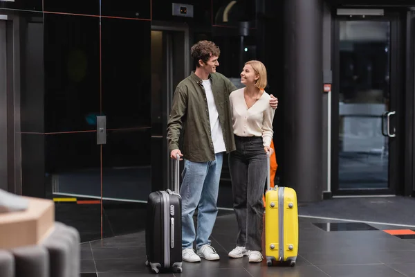 Full length of young and happy couple smiling at each other near luggage in hotel lobby - foto de stock
