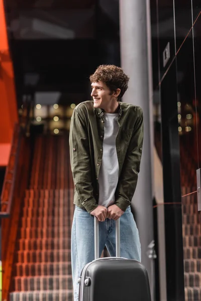 Cheerful young man with suitcase standing in hotel lobby and looking away - foto de stock