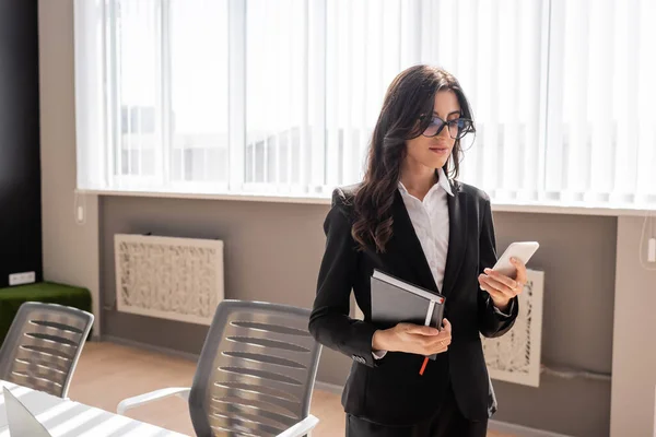 Businesswoman in eyeglasses holding notebook and messaging on mobile phone in office — Stockfoto