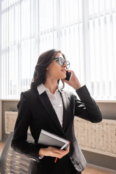 Brunette office manager in eyeglasses holding notebook and looking away while talking on mobile phone — Stock Photo