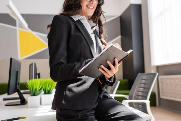 Cropped view of smiling businesswoman writing in notebook near office desk with computers — Stock Photo