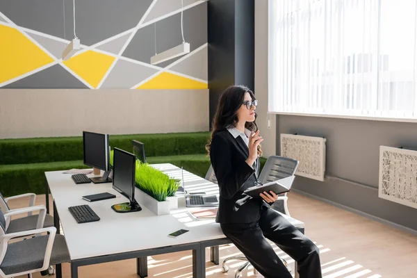 Вдумчивая деловая женщина с ноутбуком глядя в сторону стола с компьютерами в офисе — стоковое фото