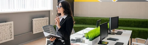 Brunette businesswoman in eyeglasses holding notebook while thinking near desk with monitors in office, banner — Stockfoto