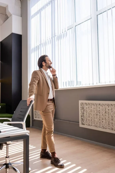 Full length of smiling businessman in beige suit touching chin and looking away near large window in office — Stockfoto