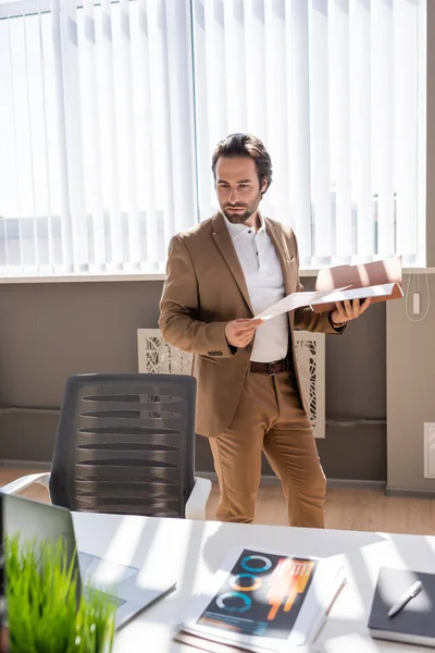 Serious businessman holding documents while looking at laptop on desk in office — Stockfoto
