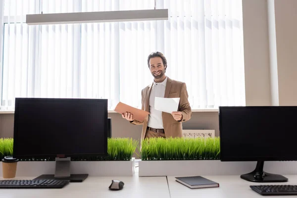 Young businessman holding documents and smiling at camera near computer monitors with blank screen in office — Stockfoto