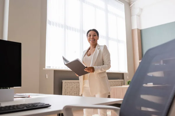 Cheerful multiracial businesswoman with folder looking at camera near work desk in office — Photo de stock