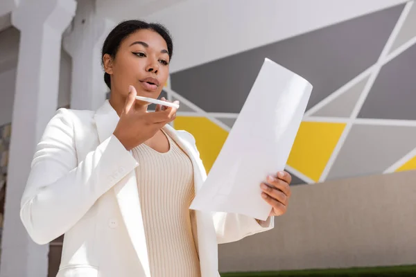 Low angle view of multiracial businesswoman looking at document and recording voice message on mobile phone — Stock Photo