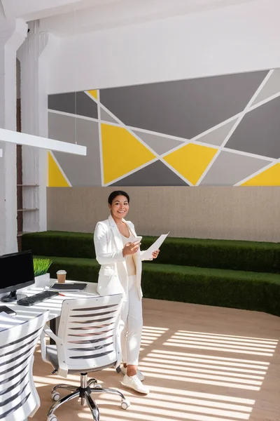 Happy multiracial businesswoman with mobile phone and paper looking at camera near desk and chairs in modern office — Fotografia de Stock