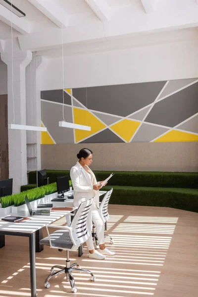 Full length of multiracial businesswoman in white suit standing with documents near desk with computers in spacious office — Foto stock