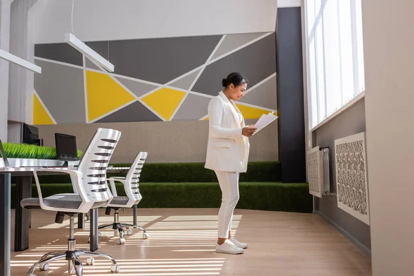 Side view of multiracial businesswoman in white suite standing with documents in office — Stock Photo