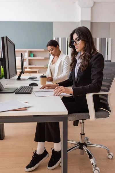 Interracial businesswomen in formal wear working with documents and computers in office — Stock Photo