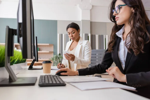 Multiracial businesswoman looking at smartphone and writing in notebook near colleague working on computer — Stockfoto